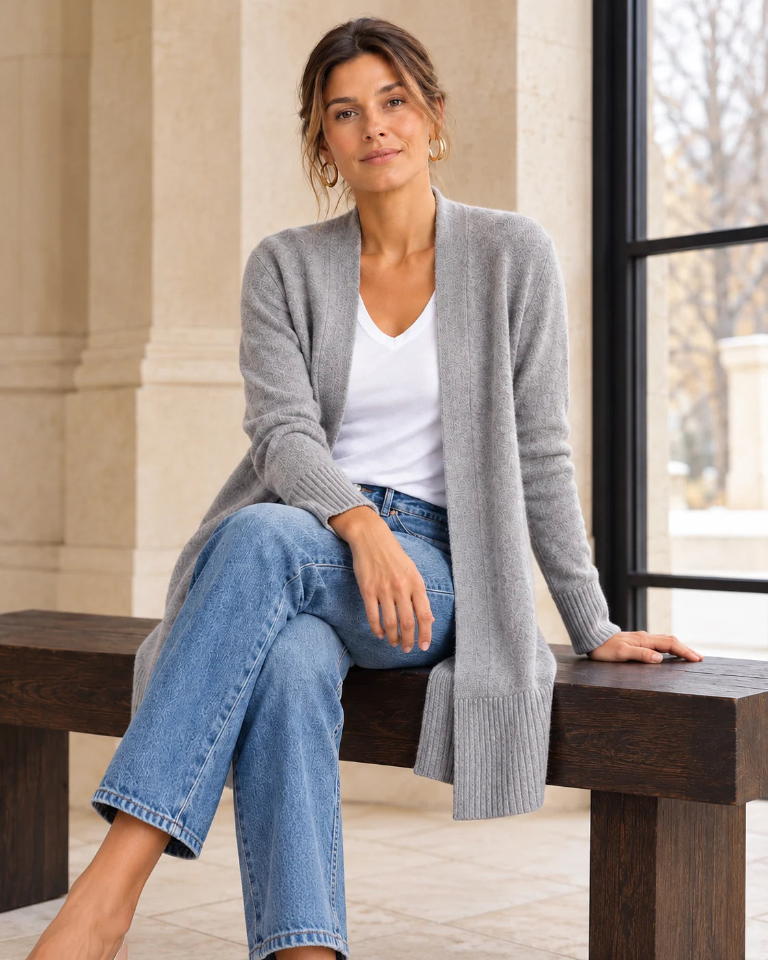 Woman in a grey cardigan and jeans, seated on a wooden bench beside a window.